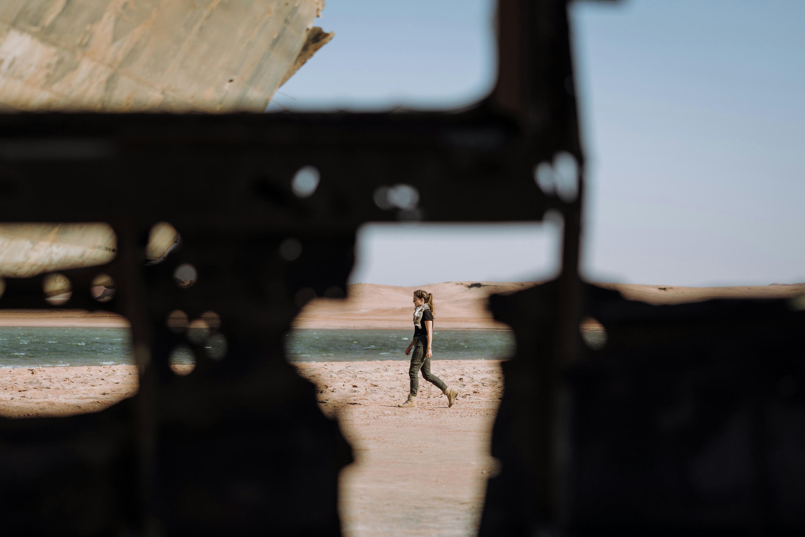 Woman viewed thru damaged metal, walks along a beach